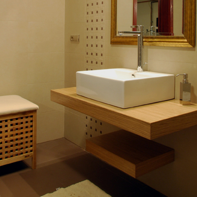 A compact bathroom vignette centers on a white rectangular basin set over a wood counter with a dark open shelf below. A chrome faucet and small dispenser stand behind the basin, and a cropped woven-front cabinet appears at the left edge. Warm tan walls, a mirror, and pale fabric cropped along the bottom edge complete the scene.