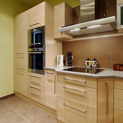 A square image shows a beige kitchen with a tiled backsplash and light-wood cabinet fronts. A built-in oven and microwave stand at left, while a white counter and sink run beneath a large stainless-steel range hood. Small kitchen appliances and containers sit on the counter against a warm yellow-green wall.