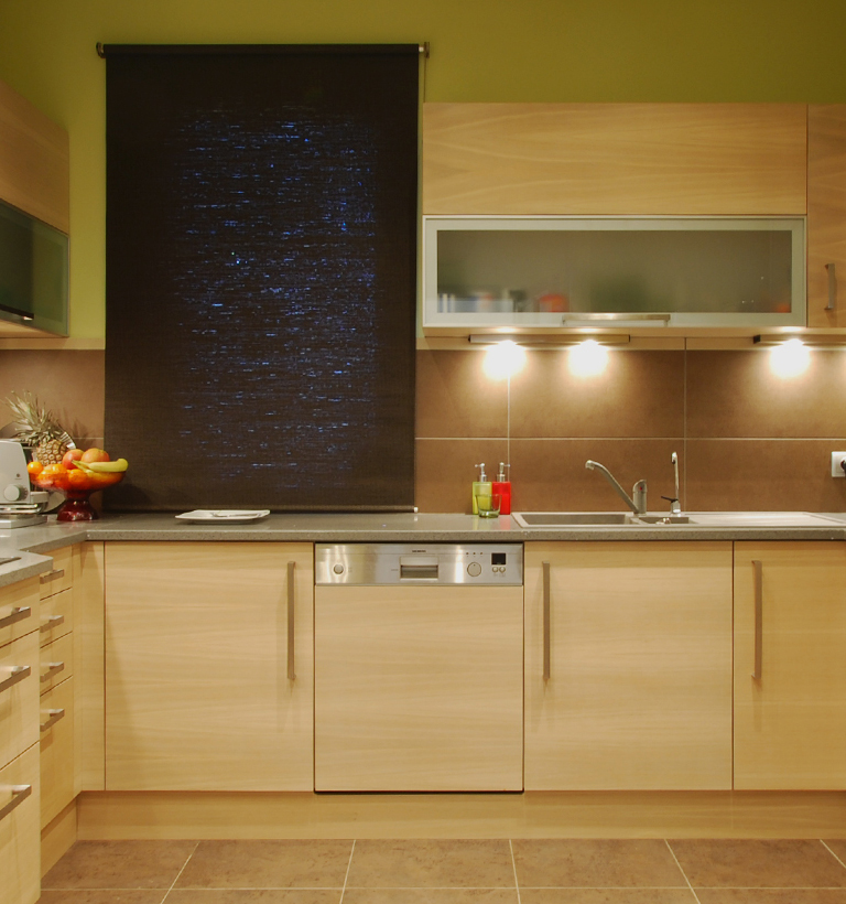 A straight-on view shows a warm beige kitchen with wood-tone lower cabinets and matching upper cabinets. A tall dark rectangular panel sits above the counter near the center-left, and a broad glass-front cabinet is centered above a sink and curved faucet. A metal-front appliance with a small display and buttons sits beneath the worktop. A bowl of colorful fruit and a small plant appear at the far left.