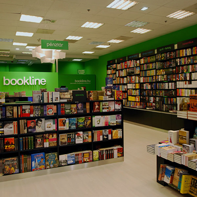 The interior shows long display counters stocked with books and tall wall shelves densely filled with book spines at the back. Bright green wall panels carry a large “bookline” sign, and a smaller overhead sign reads “pénztár”. Rectangular ceiling lights run in rows above the counters.