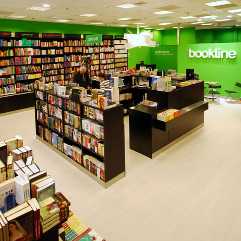 A bookstore interior is arranged with long black book racks, wall shelves, and checkout counters stocked with colorful books. Two people are visible among the displays near the middle-left. Bright green walls carry large white “bookline” lettering and smaller “bookline.hu” text. Ceiling lights illuminate the pale floor and the dense rows of books.