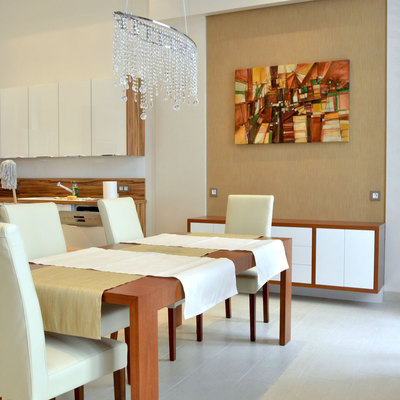 A bright dining room contains a rectangular table covered with a white cloth and surrounded by chairs with pale upholstered backs. A chandelier hangs above, and low white panels run along the back wall beneath a framed picture. The neutral room has light walls, a pale floor, and a tall glazed opening at right.