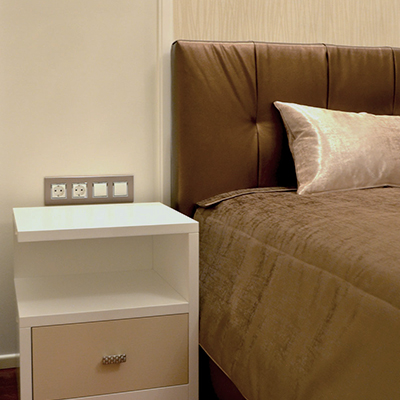 A bed with a brown quilted cover and a tall segmented brown headboard fills the right side of the image. A pale rectangular pillow rests near the headboard, and the cover drapes across the mattress. A white bedside cabinet with a beige drawer and small metal handle sits at left below a row of wall switches.