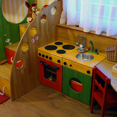 A colorful toy kitchen has a yellow countertop with black circular burners, a metal-look faucet, and a round sink. Red, green, and wood-colored cabinet doors form an oven and storage fronts below the counter. A small toy figure is visible behind an upright wood-colored panel, and a wicker basket sits on the counter at the far right.