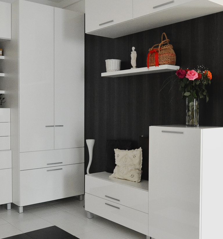 A dark charcoal wall and white cabinetry frame a narrow kitchen display. A white shelf holds a small white vase, a figurine, and a woven basket with red ribbon, while a glass vase of red, pink, and orange flowers stands on the counter. A pale patterned cushion rests on a low drawer beside a tall white vase. The close vertical composition is bordered by pale cabinet fronts.