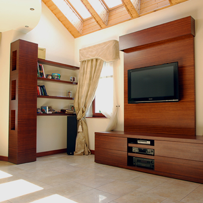 Dark wood built-in shelving and cabinetry line the left and right sides of a bright room, with books and small objects on the shelves. A television is mounted on a broad wood panel above media shelves and drawers at the right. A light curtain hangs beside a window below a sloped ceiling with exposed timber beams.