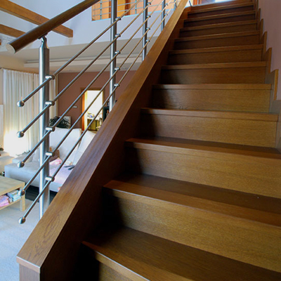 Dark wood steps rise from the foreground to the upper right, bordered by a broad wood handrail. A silver metal railing with horizontal bars follows the open side of the staircase. A light-colored room and curtained window are visible through the railing.
