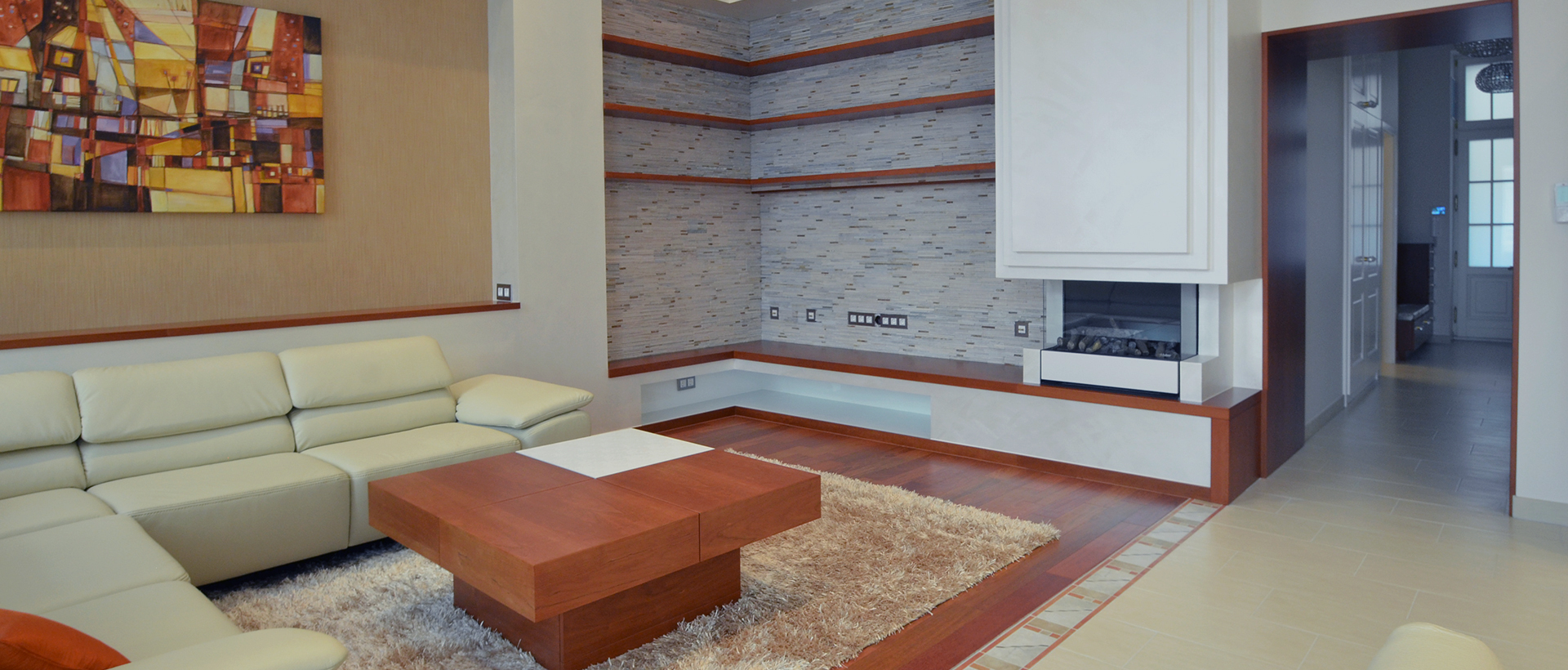 A wide living-room view shows a pale sectional sofa along the left wall and a low wooden coffee table on a cream shag rug. Behind the seating, a gray tiled feature wall with orange-brown bands surrounds a dark rectangular opening. A colorful abstract picture hangs at the upper left, and a doorway at right opens into a dimmer adjoining space. The room combines pale upholstery and walls with warm wood and darker accents.
