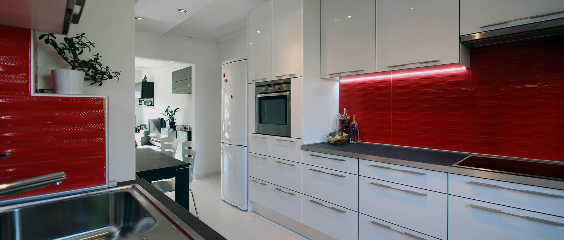 A wide kitchen view centers on glossy red cabinetry and a long bank of pale drawers beneath a dark countertop. A black glass cooktop fills the foreground island, while a leafy plant in a white pot sits on the counter at upper left. Pale upper cabinets span the room, with an oven and dining chairs visible toward the back. The red and white cabinetry contrasts with the dark work surfaces.
