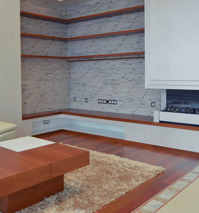 A cropped living-room view shows a corner wall covered in small gray rectangular tiles crossed by narrow orange-brown bands. A white wall unit occupies the upper right above a dark rectangular recess with small objects inside. A cream shag rug lies across the wood floor beside the corner of a brown wooden coffee table, while part of a pale upholstered sofa enters from the left. The image focuses on the floor and tiled wall rather than the full room.