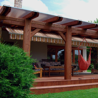 A timber pergola covers a raised wooden deck, with thick posts and crossbeams framing the scene. A red fabric hanging swing seat hangs near the right side beneath the beams. A striped awning spans the shaded area behind a wooden bench, and a leafy green shrub fills the left foreground.