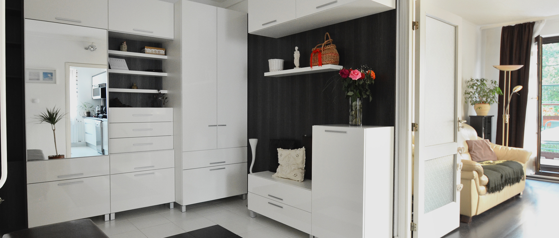 Tall white cabinets and drawers fill the left side of a bright interior, with a dark recessed display shelf near the center. A potted plant stands at the left, and an open white doorway reveals a pale sofa with cushions, a floor lamp, dark curtains, and a bright window. The recess contains a basket, small ornaments, and a vase of flowers. The wide composition links the cabinetry with the adjoining sitting area.