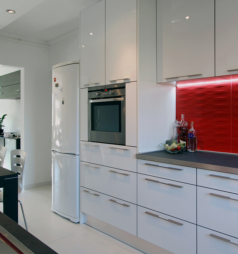A modern kitchen has white cabinets, a tall refrigerator at left, and a dark built-in oven near the center. Glossy red cabinet panels line the counter on the right, where bottles and a bowl sit beside the sink. A dark dining table edge and pale chairs enter the lower-left foreground. The room is shown in a wide, slightly angled view.