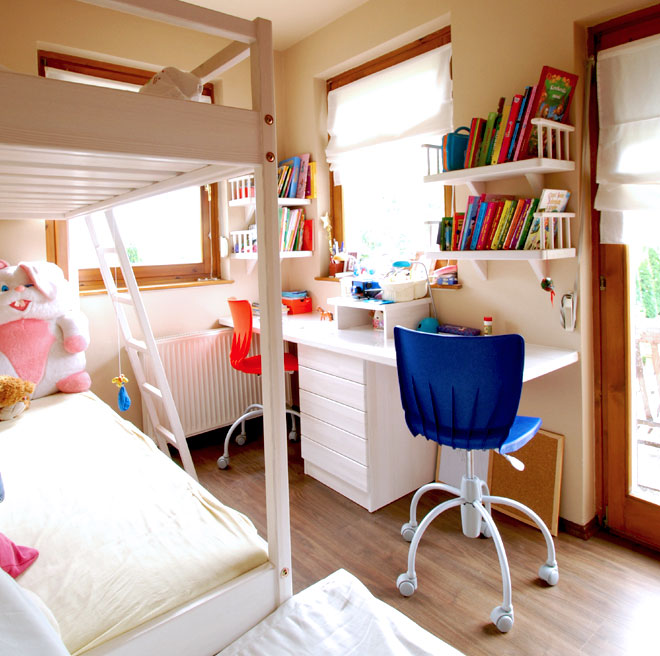 A white bed frame with an elevated rail fills the left foreground, with a white-covered mattress below it. A compact white desk with drawers sits beneath shelves lined with colorful books, with a blue office chair in front. Bright windows frame the back and right sides, and part of a large plush rabbit is visible at the far left.