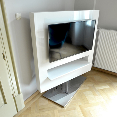 A white entertainment unit surrounds a flat-screen television and includes a long open shelf below it. The unit stands above a gray glass-topped metal base on a light patterned wood floor. A white radiator is mounted on the wall to the right beside pale gray walls.