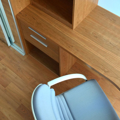 A wood-grain desk wraps around a corner, with a broad work surface and two narrow drawers beneath its near section. A pale blue chair with curved arms sits in front, partially cropped by the lower edge. A narrow window is visible beside the desk, and matching wood flooring continues across the room.
