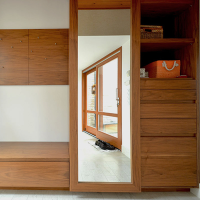 A fitted wood-grain storage unit spans the view, with cupboard fronts and wide drawers at the left and right. A full-height central mirror reflects a light tiled area and an orange-brown glazed door, with dark footwear near the threshold. Open shelves at the right hold a woven basket and an orange bag with white handles.