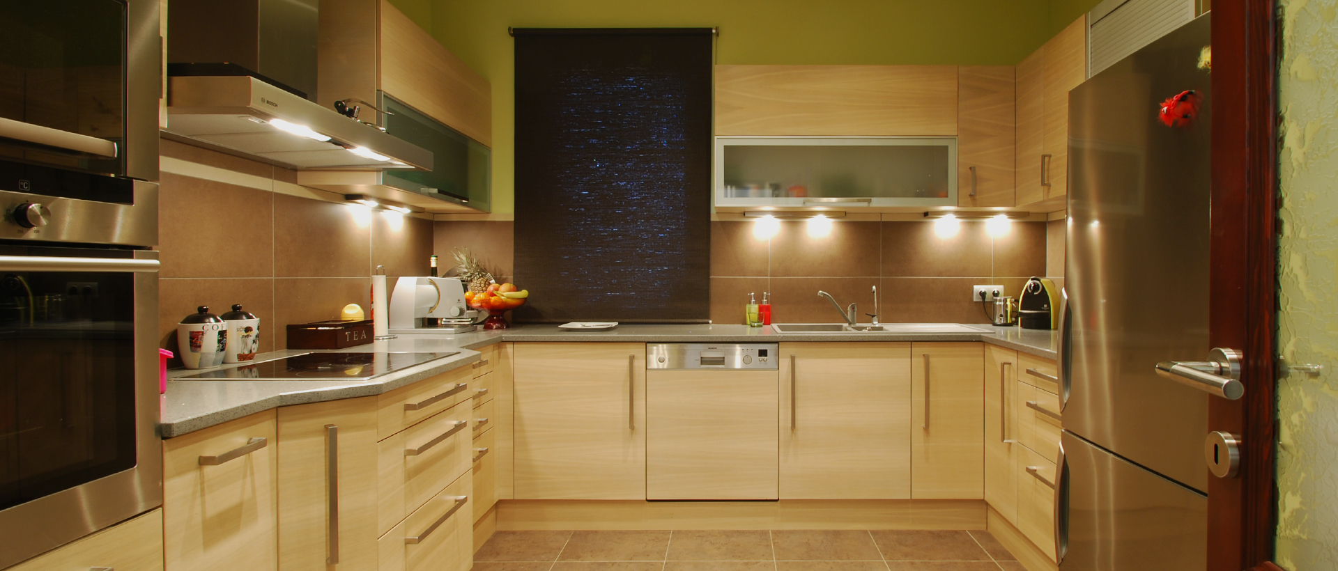 An L-shaped wood-tone kitchen fills the frame, with pale countertops and matching cabinet fronts running along the back and side walls. A tall dark rectangular panel hangs above the central counter, where a sink, curved faucet, bottles, and small appliances are arranged. A broad black-front refrigerator stands at the right, while a range hood and dark appliance are visible at the left. Under-cabinet lights illuminate the work surfaces.