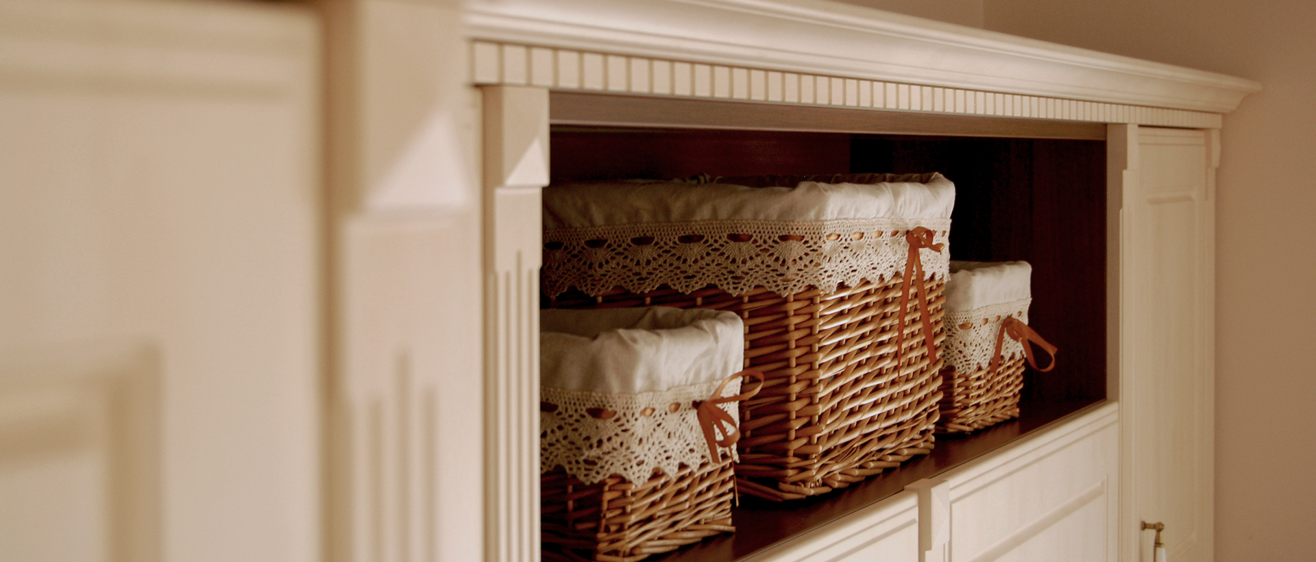 A recessed display niche contains several woven brown baskets lined with pale fabric and edged with white crochet trim. The baskets sit on dark shelves within a broad white cabinet frame. A pale wall fills the left side, and part of a white cabinet door or panel frames the right. This wider view shows the basket display built into the surrounding cabinetry.