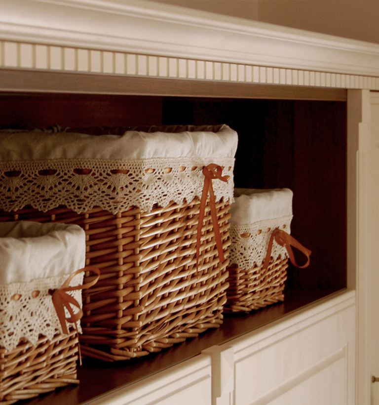 Several rectangular wicker baskets with pale fabric liners and crocheted white trim are arranged on dark display shelves. The baskets have woven brown sides and visible loop handles. White cabinet framing surrounds the recessed display. The close composition emphasizes the baskets and their contrasting woven and fabric textures.